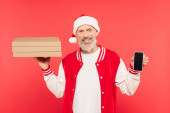 šťastný muž středního věku v Santa klobouk drží krabice od pizzy a smartphone s prázdnou obrazovkou izolované na červené
