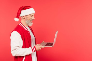 Noel Baba şapkalı neşeli orta yaşlı serbest çalışanın yan görüntüsü kırmızı ekranda izole edilmiş bir dizüstü bilgisayar.