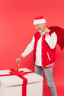 Şapkalı neşeli, orta yaşlı bir adam Noel Baba torbasını tutuyor ve kırmızı üzerine izole edilmiş büyük hediyelere bakıyor.