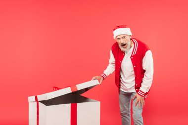 Şok olmuş orta yaşlı Noel Baba şapkalı adam kırmızı üzerine izole edilmiş büyük bir hediye açıyor.
