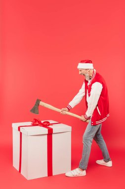 full length of middle aged man in santa hat holding axe while screaming near huge present on red clipart