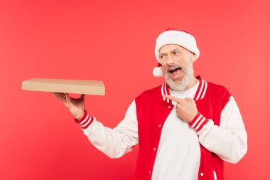 Noel Baba şapkalı orta yaşlı adam kırmızı üzerine izole edilmiş pizza kutusunu işaret ediyor.