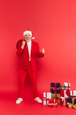 Şapkalı ve takım elbiseli mutlu orta yaşlı adam hediyelerin yanında Noel Baba çuvalı tutarken kırmızıda başparmağını gösteriyor.