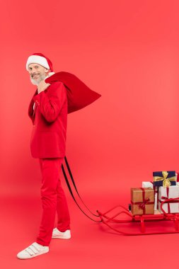 Noel Baba şapkalı ve takım elbiseli neşeli orta yaşlı bir adam elinde hediyeler ve kırmızı çuvalla kızak tutuyor.