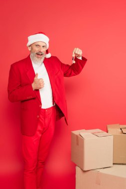 Orta yaşlı takım elbiseli ve Noel Baba şapkalı bir adam anahtarı tutuyor ve kırmızı kutuların yanında baş parmağını gösteriyor.