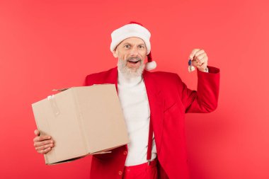 Heyecanlı orta yaşlı takım elbiseli ve Noel Baba şapkalı adam elinde karton kutu ve anahtarı kırmızı üzerine izole edilmiş.