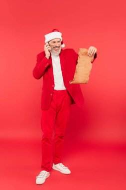 Tam boy, Noel Baba şapkalı olgun bir adam el sanatları kağıdında mektup okurken kırmızıda akıllı telefonla konuşuyor.