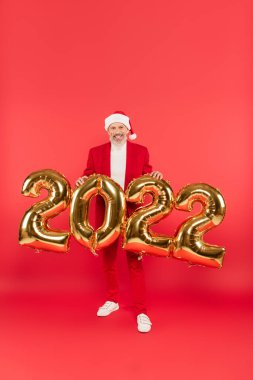 Noel Baba şapkalı mutlu bir adam 2022 balonlarının yanında kırmızı üzerinde duruyor.