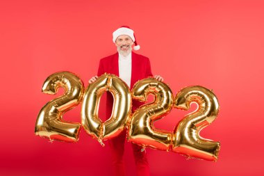 Gülümseyen, Noel Baba şapkalı olgun bir adam 2022 balonlarının yanında kırmızı üzerinde duruyor.