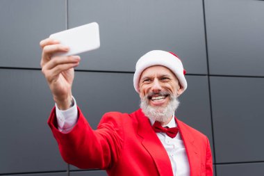 Noel Baba şapkalı mutlu adam gri duvarın yanında selfie çekiyor.