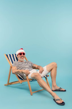 Noel Baba şapkalı ve güneş gözlüklü mutlu bir adam mavi sandalyede oturuyor.