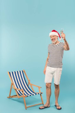 Noel Baba şapkalı olgun bir adam elinde mavi şampanyayla güvertedeki sandalyenin yanında.