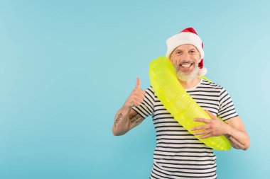 Pozitif orta yaşlı, Noel Baba şapkalı, elinde şişme yüzük tutan bir adam mavi üzerinde parmağını gösteriyor.