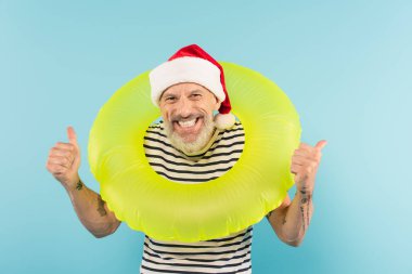 Pozitif orta yaşlı, Noel Baba şapkalı, elinde şişirilebilir bir yüzük tutan bir adam mavi üzerinde parmaklarını gösteriyor.