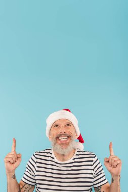 Dikenli tişörtlü ve Noel Baba şapkalı neşeli orta yaşlı bir adam mavi üzerinde parmakları olan birini işaret ediyor.