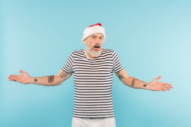 Dikenli tişört ve Noel Baba şapkalı kafası karışmış orta yaşlı bir adam mavi üzerine izole edilmiş.