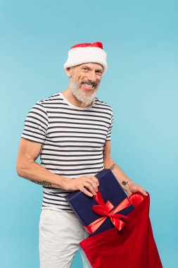 Noel Baba şapkalı mutlu orta yaşlı adam mavi bir çuvala saklanmış hediyesini tutuyor.