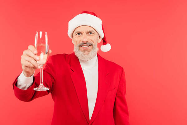 счастливый мужчина средних лет в костюме и шляпе Санты держа бокал шампанского изолированы на красный