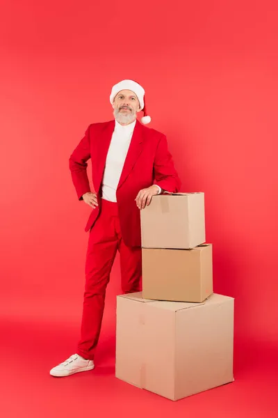 middle aged man in suit and santa hat leaning on carton boxes on red