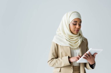 young muslim woman in hijab using digital tablet isolated on grey clipart