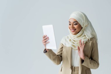 tesettürlü mutlu Müslüman kadın dijital tablet tutuyor ve el sallıyor. Video çağrısı gri üzerine izole edilmiş.
