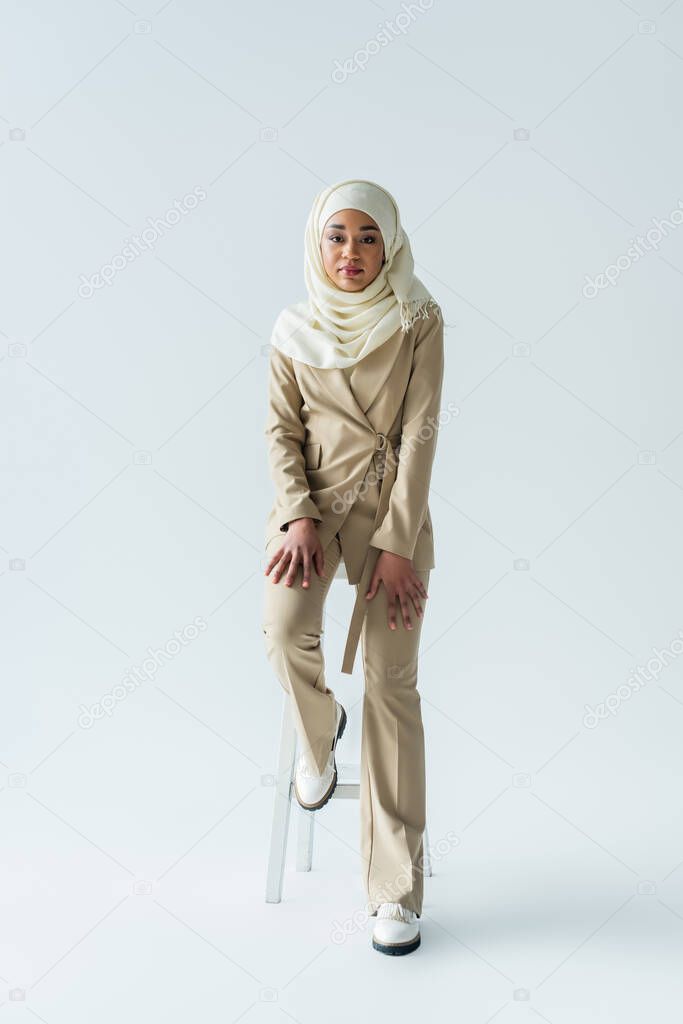 full length of stylish muslim woman in hijab sitting on stool on grey 