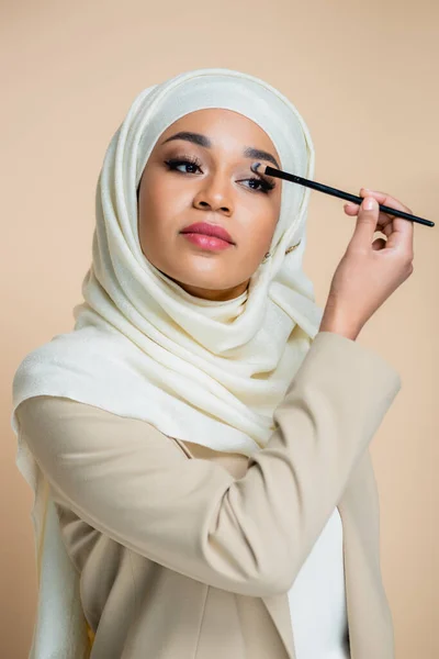 young muslim woman in hijab applying eye shadow isolated on beige