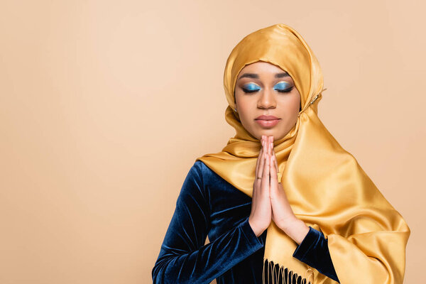 young muslim woman in hijab standing with praying hands isolated on beige