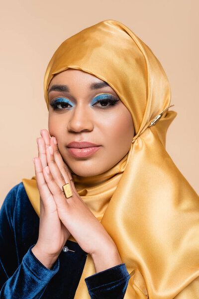 muslim woman in yellow hijab with bright makeup and golden ring looking at camera isolated on beige