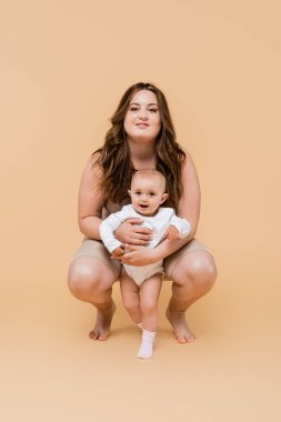 Happy plus size mom hugging baby daughter on beige background  clipart