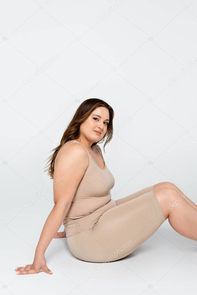 Smiling young body positive woman looking at camera while sitting on grey background