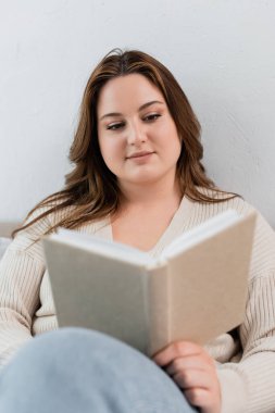 Aşırı kilolu esmer bir kadın evde bulanık bir kitap okuyor. 