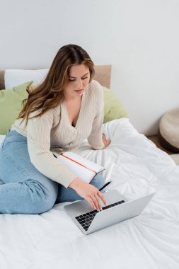 Yatağın yanında dizüstü bilgisayar kullanan genç ve serbest çalışan biri var. 