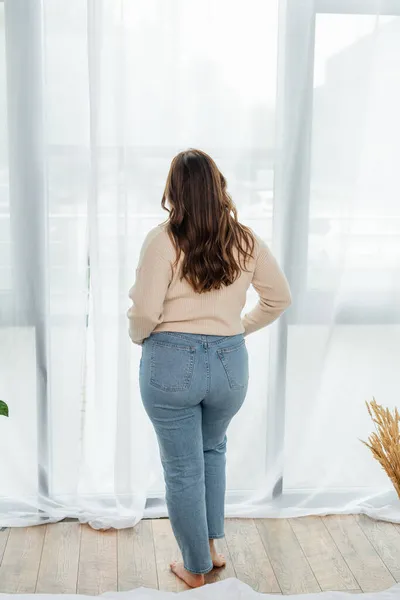 Back view of body positive woman standing near window at home