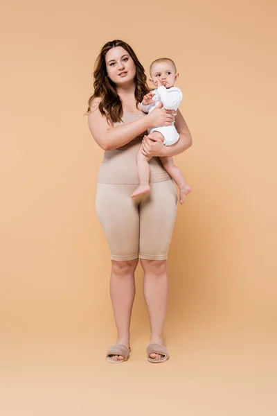 Full length of body positive mother holding baby daughter on beige background