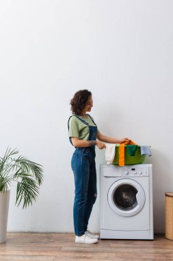 Çamaşır makinesinin üstünde çamaşır kâsesinin yanında duran kot pantolonlu ev kadını.