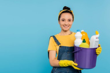 Sarı lastik eldivenli genç bir ev hanımı. Elinde kovayla mavi renkli deterjanlar var.