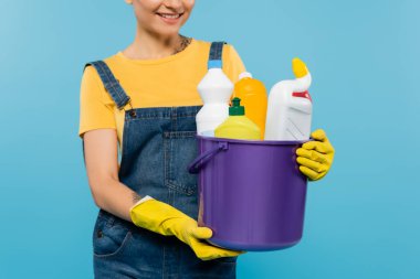 Sarı lastik eldivenli gülümseyen ev kadınının mavi üzerine izole edilmiş temizleyicileri olan kovayı tutuşu.