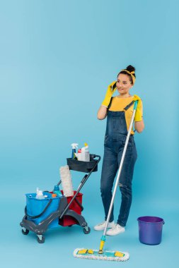 Paspaslı temizlikçi akıllı telefondan konuşuyor, alışveriş arabasının yanında, temizleme malzemeleri mavi üzerine.