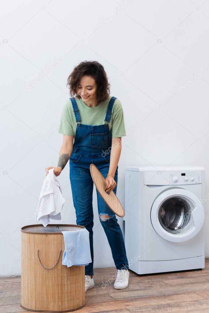 smiling woman holding laundry near basket and washing machine
