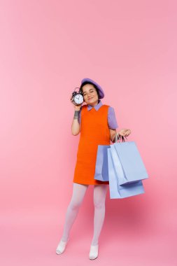 Kapalı gözlü, elinde klasik çalar saat ve pembe renkli alışveriş torbaları olan mutlu bir kadın.