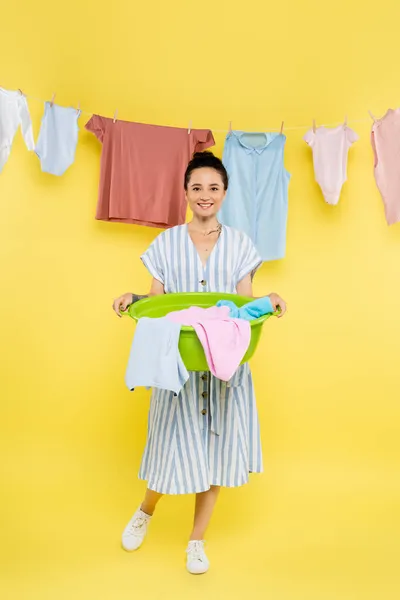 微笑的家庭主妇在黄色背景上挂着的衣服旁边拿着洗衣碗 — 图库照片