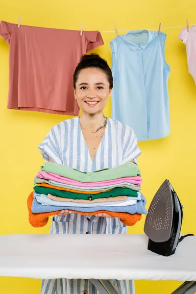 Mujer Alegre Sosteniendo Pila Ropa Cerca Plancha Tabla Planchar Amarillo —  Fotos de Stock