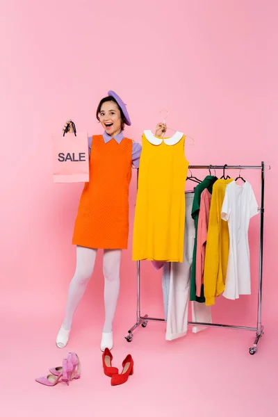 靴の近くに販売ショッピングバッグと黄色の袖のないドレスとピンクの服を着たラックを持つ驚きの女性 — ストック写真