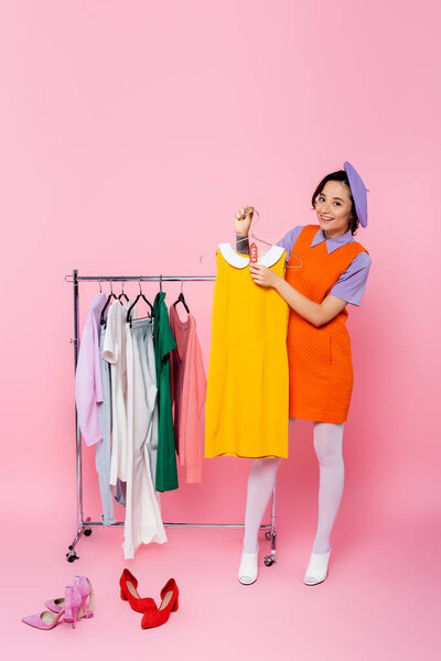 happy woman holding yellow sleeveless dress with sale tag near footwear and rack with garments on pink