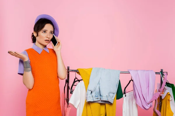 Zdenerwowana Kobieta Rozmawia Smartfonie Pobliżu Rack Ubrania Izolowane Różowy — Zdjęcie stockowe