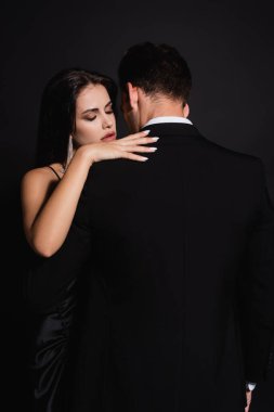 back view of man in suit near elegant woman hugging him isolated on black clipart