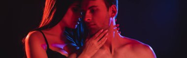 Genç ve seksi kadın, kırmızı ışıklandırmalı üstsüz adamı kucaklıyor. Siyah pankartta izole edilmiş.