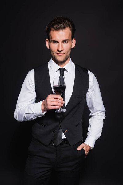 front view of smiling and elegant man holding glass of red wine while standing with hand in pocket isolated on black