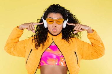 curly african american young woman in sunglasses pointing at headphones and pouting lips isolated on yellow clipart